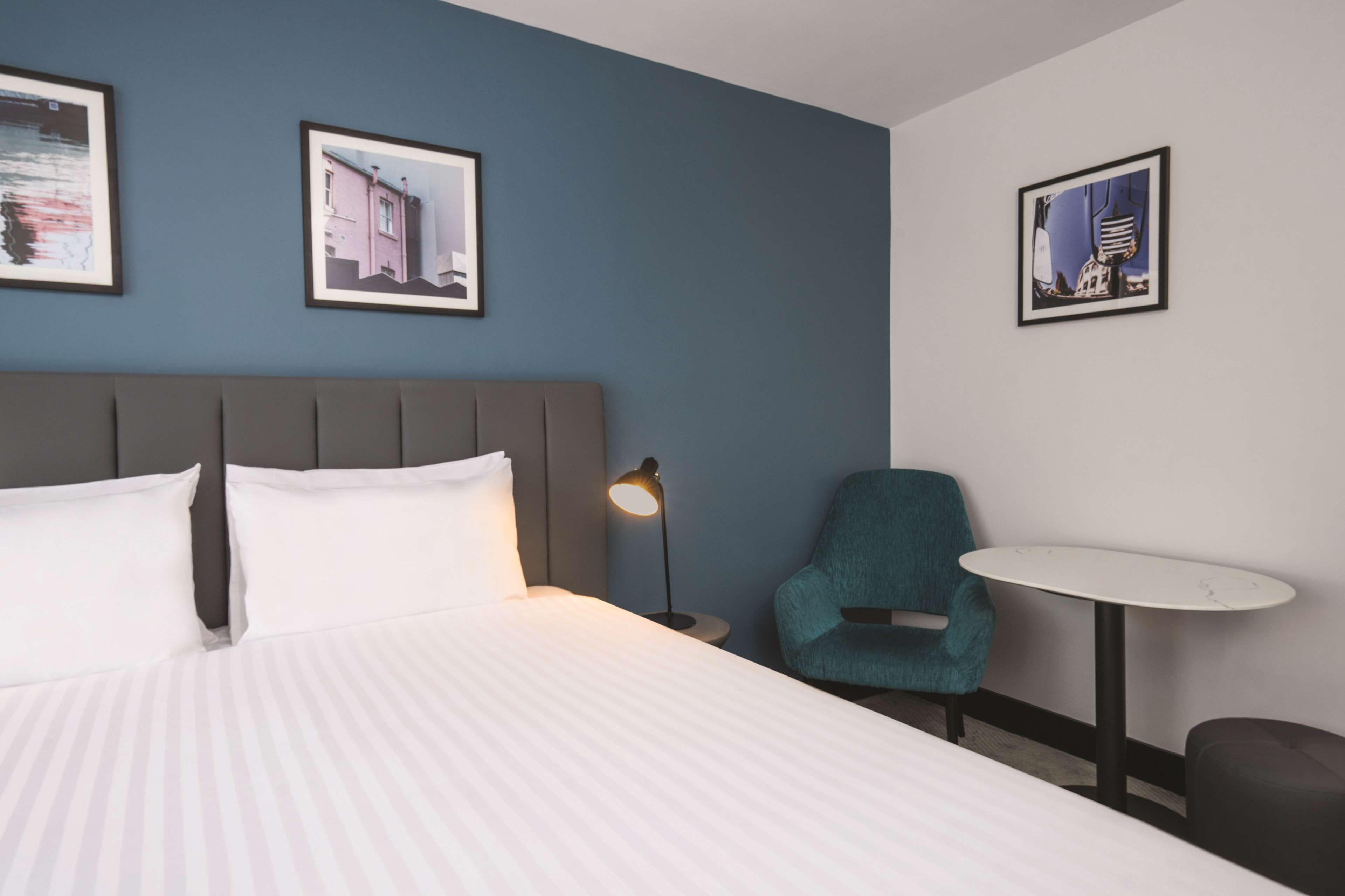 Quarto de hotel com uma cama, uma cadeira, uma mesinha e três quadros na parede.