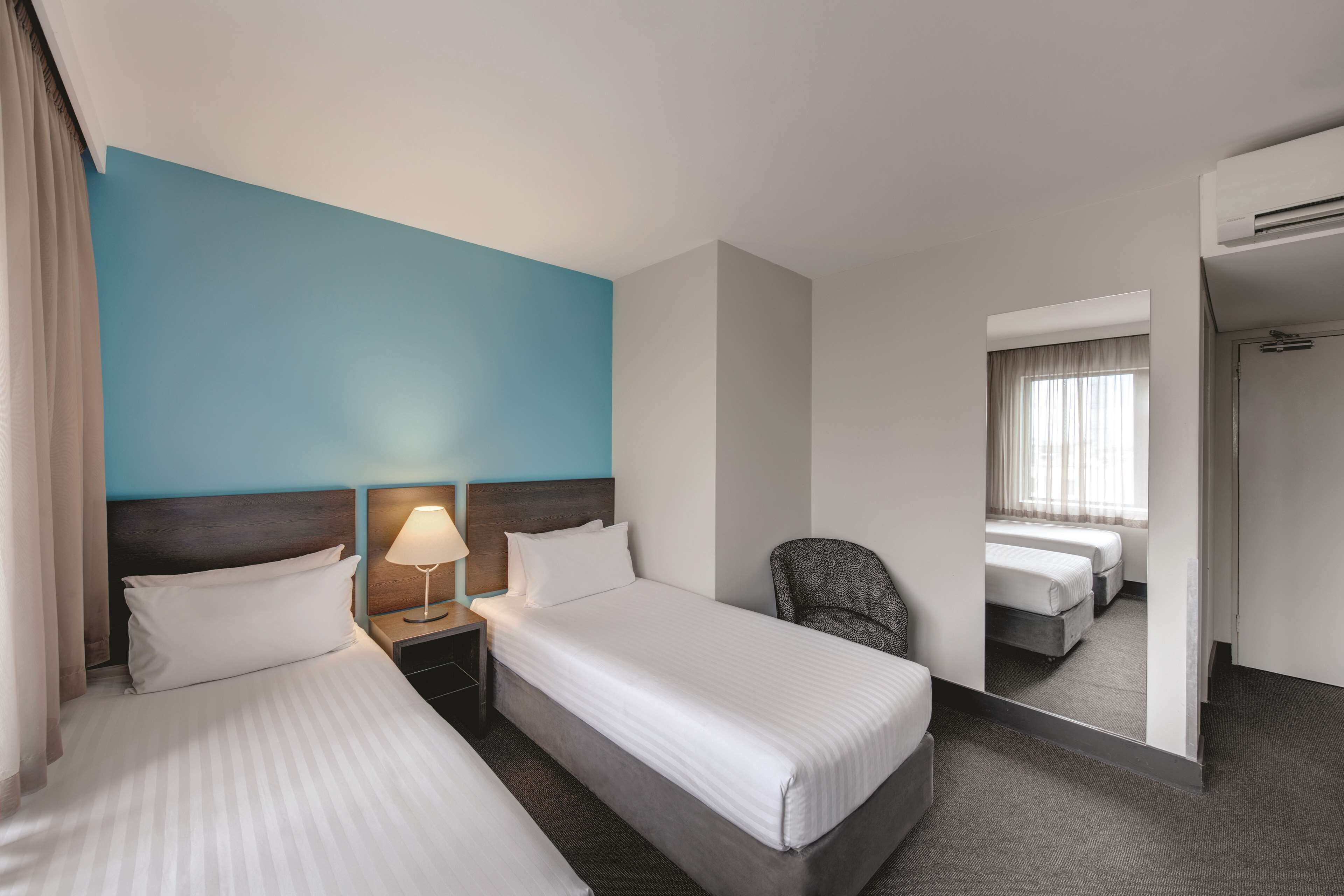 A hotel room with two beds, a nightstand with a lamp, a chair, and a mirror reflecting another room.