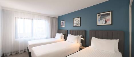 A hotel room with two beds, a large window with sheer curtains, and artwork on the walls.