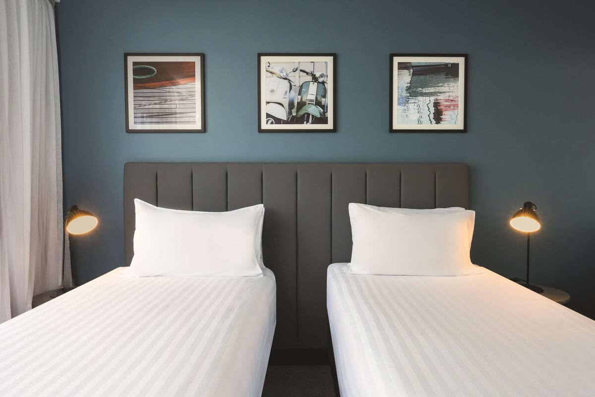 Ein Hotelzimmer mit zwei Betten, einem Kopfteil, gerahmten Kunstwerken an der Wand und Nachttischlampen.