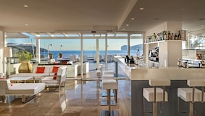 Un espace bar moderne avec vue sur la mer, doté de mobilier blanc et de différentes bouteilles exposées sur des étagères.