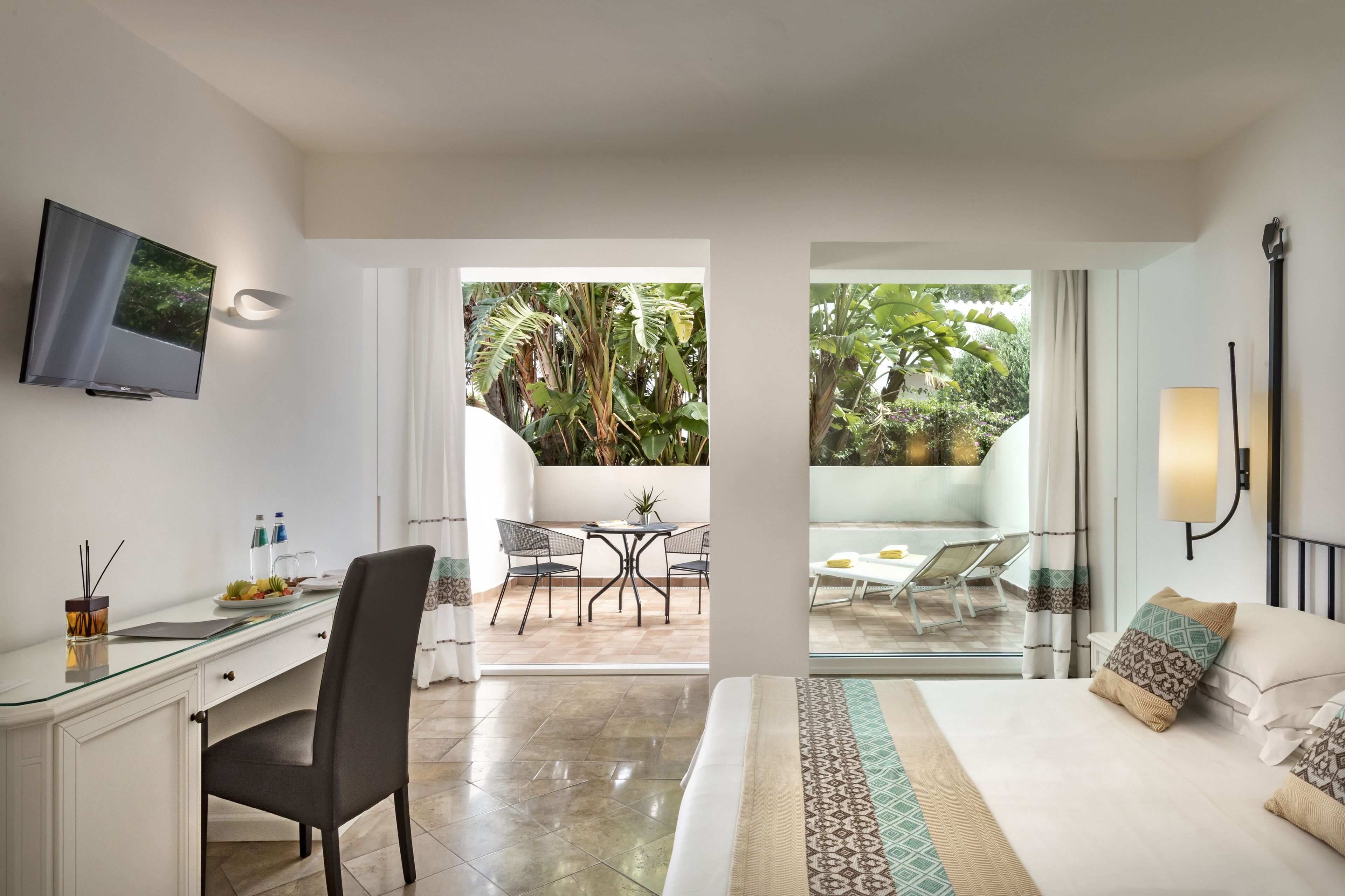 A hotel room with a desk, chair, bed, and a view of the outdoor patio area with chairs and plants.