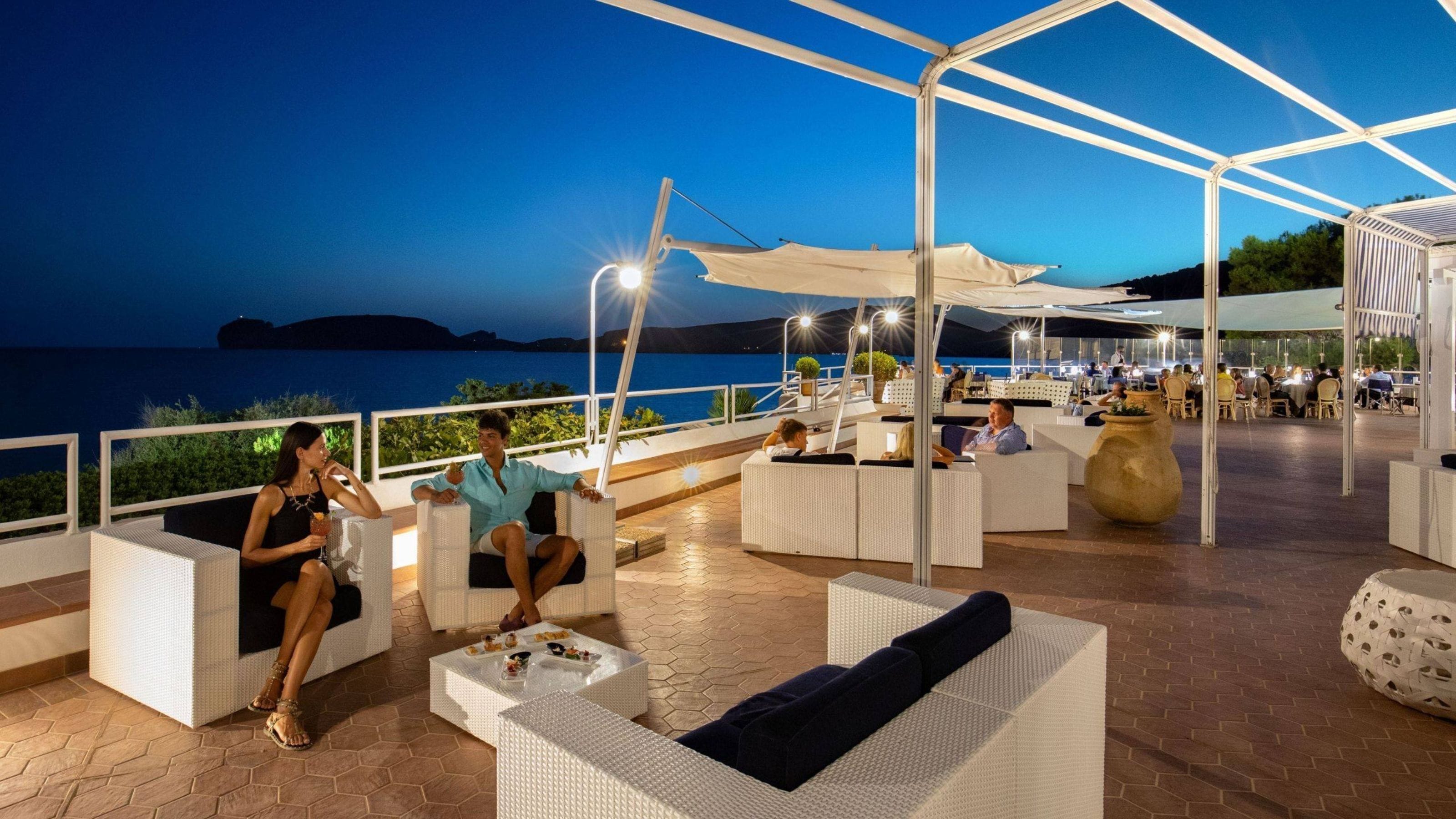 Un espace extérieur aménagé avec des meubles en osier blanc, un grand bac à fleurs et une vue sur la mer au crépuscule.