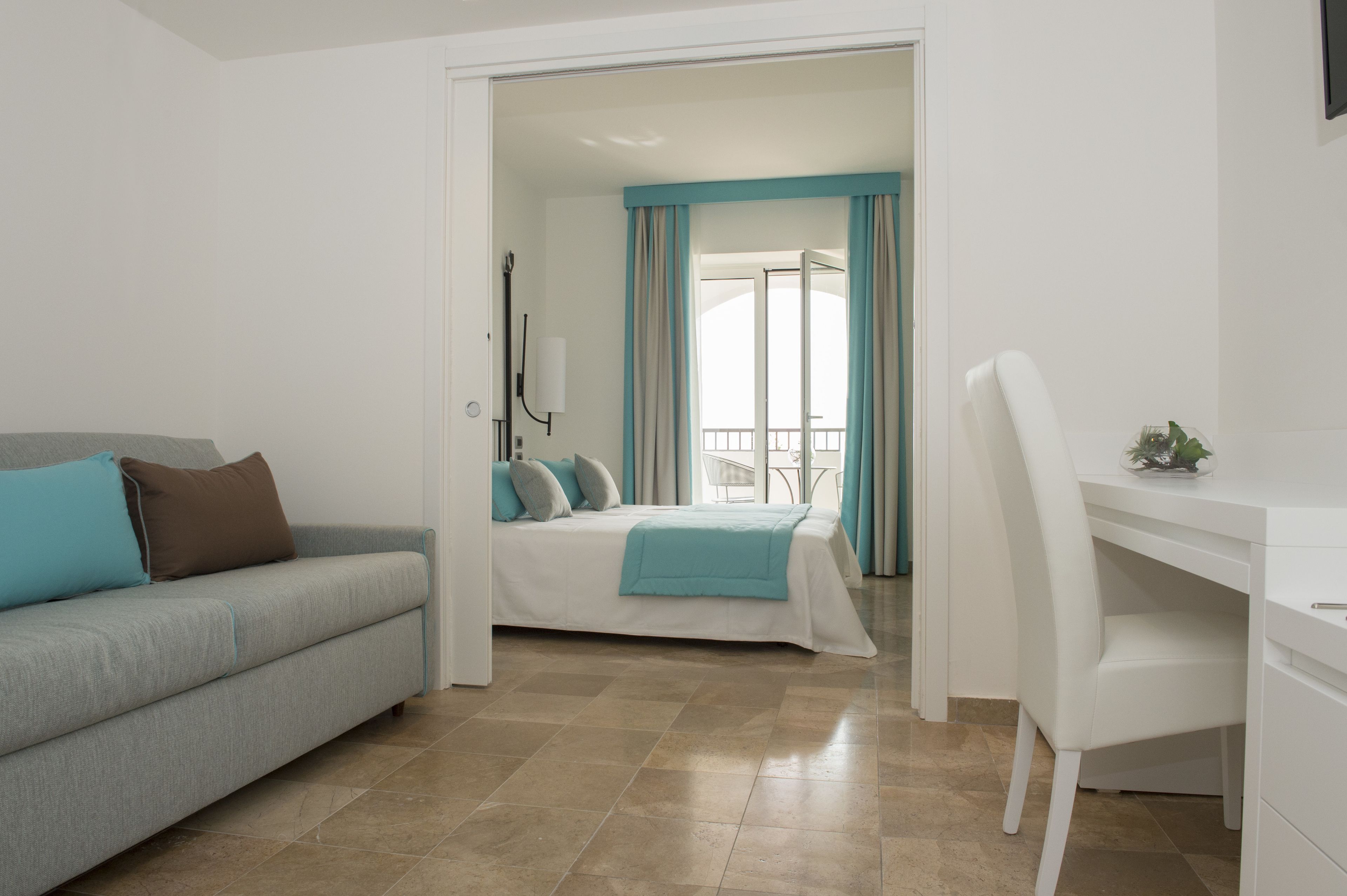 Une chambre d’hôtel moderne avec un canapé gris, un bureau blanc, un lit recouvert d’une couverture bleue et un balcon avec des rideaux.