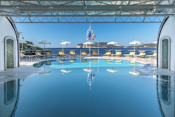 A pool area with clear blue water, lounge chairs, umbrellas, and flags. - El Faro Hotel & Spa (Alghero)