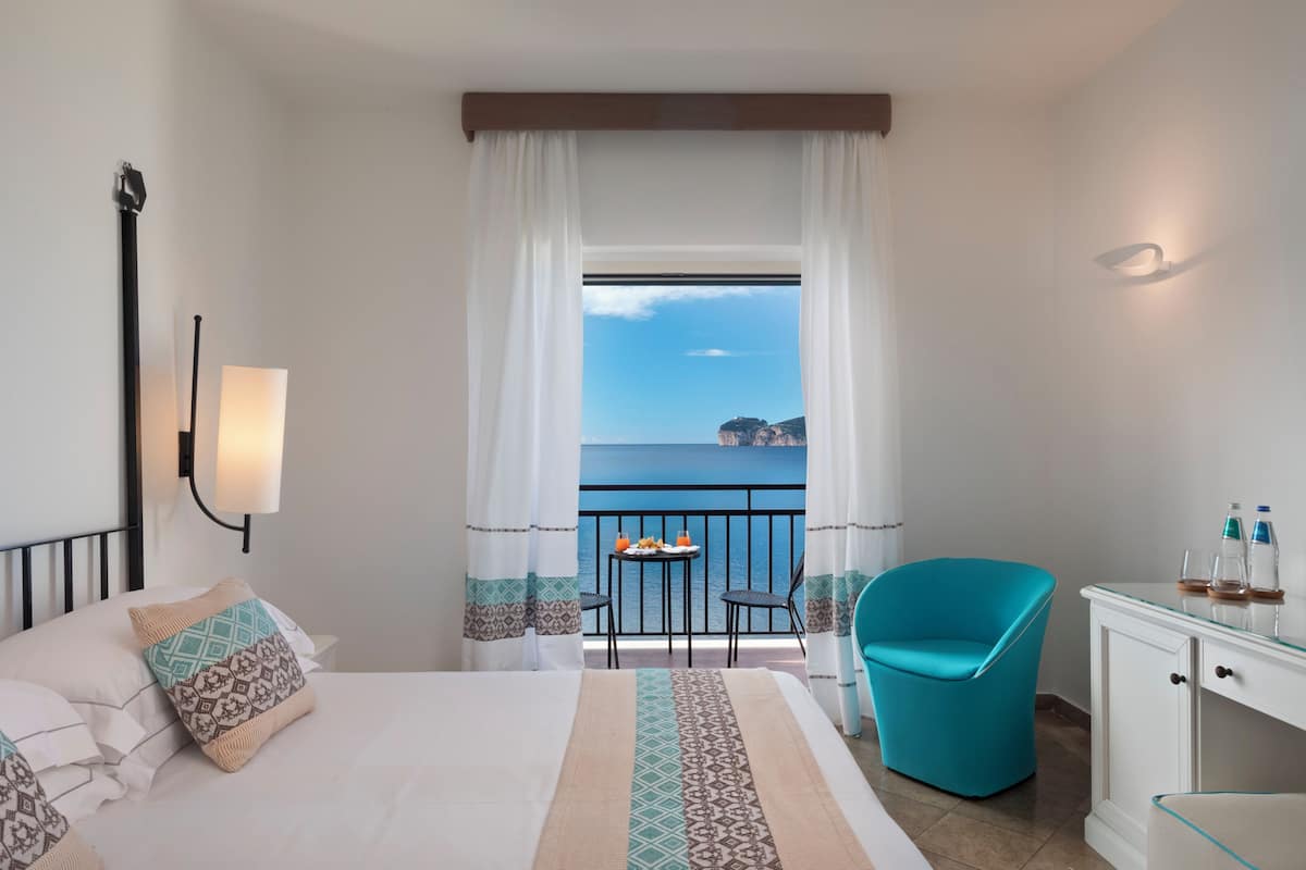 Habitación de hotel con cama, escritorio con silla, una mesa con bebidas y un balcón con vista al mar.