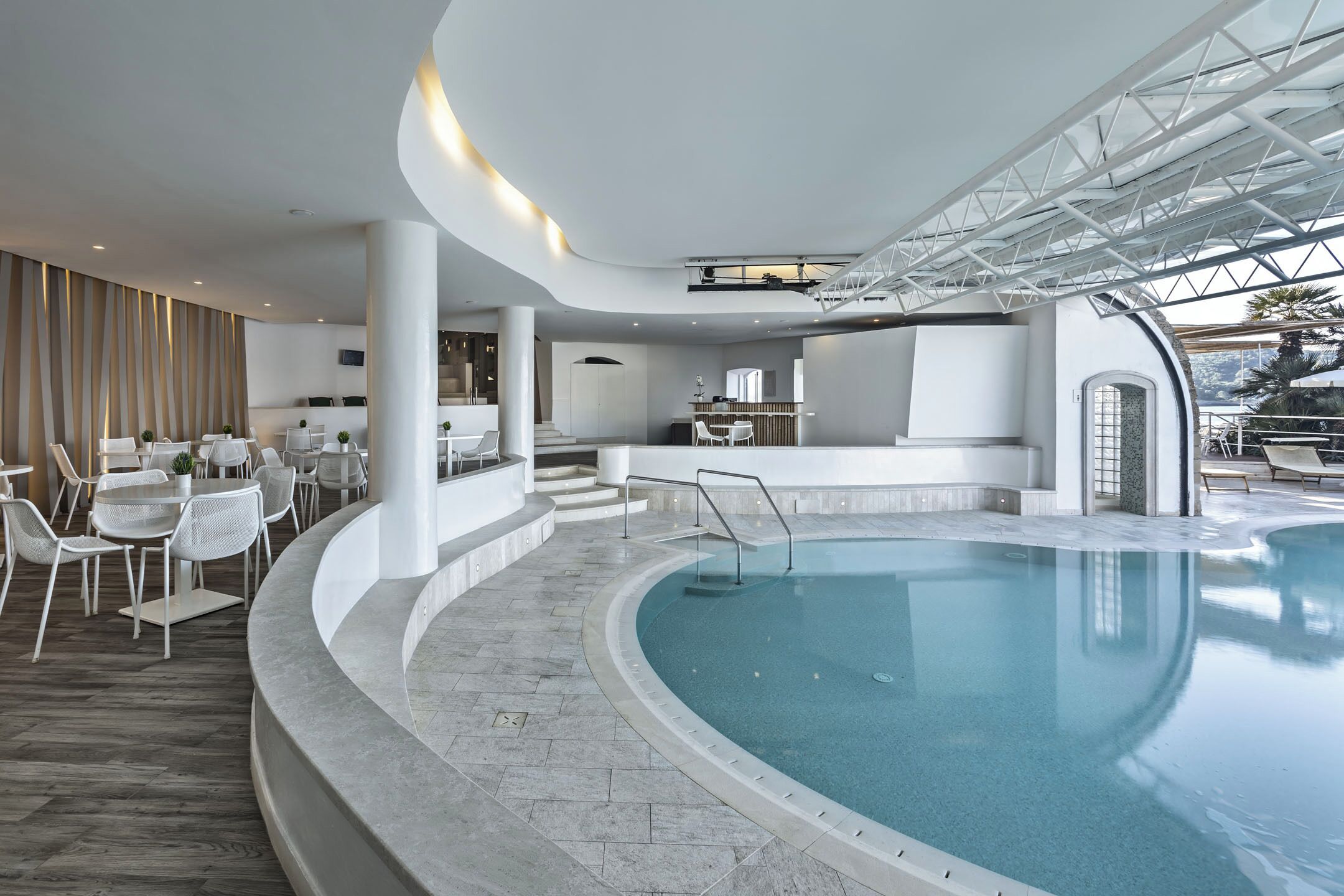 A modern indoor swimming pool with a curved design, surrounded by white furniture and a bar area.
