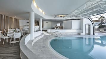A modern indoor swimming pool with a curved design, surrounded by white furniture and a bar area.