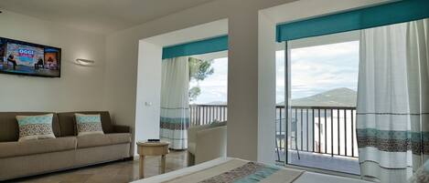 A hotel room with a bed, a sofa, a TV, a balcony with a view of the sea, and a small table.