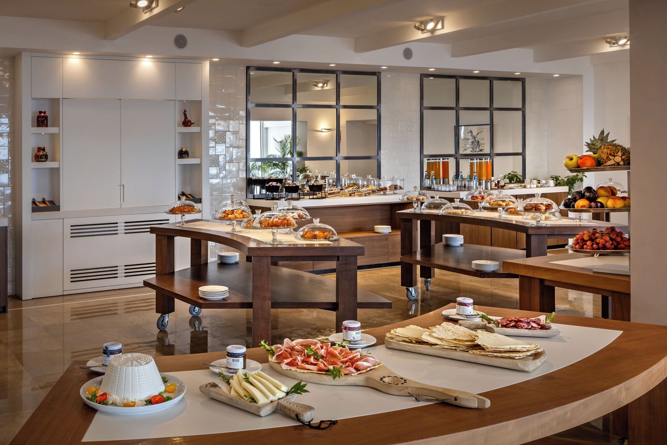 Ein moderner Hotel-Speisesaal mit mehreren Tischen, verschiedenen Speisenbuffets und einem großen Fenster, das für natürliches Licht sorgt.