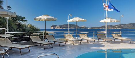 Zona de piscina con sillones de descanso y sombrillas, una bandera y vista al mar.
