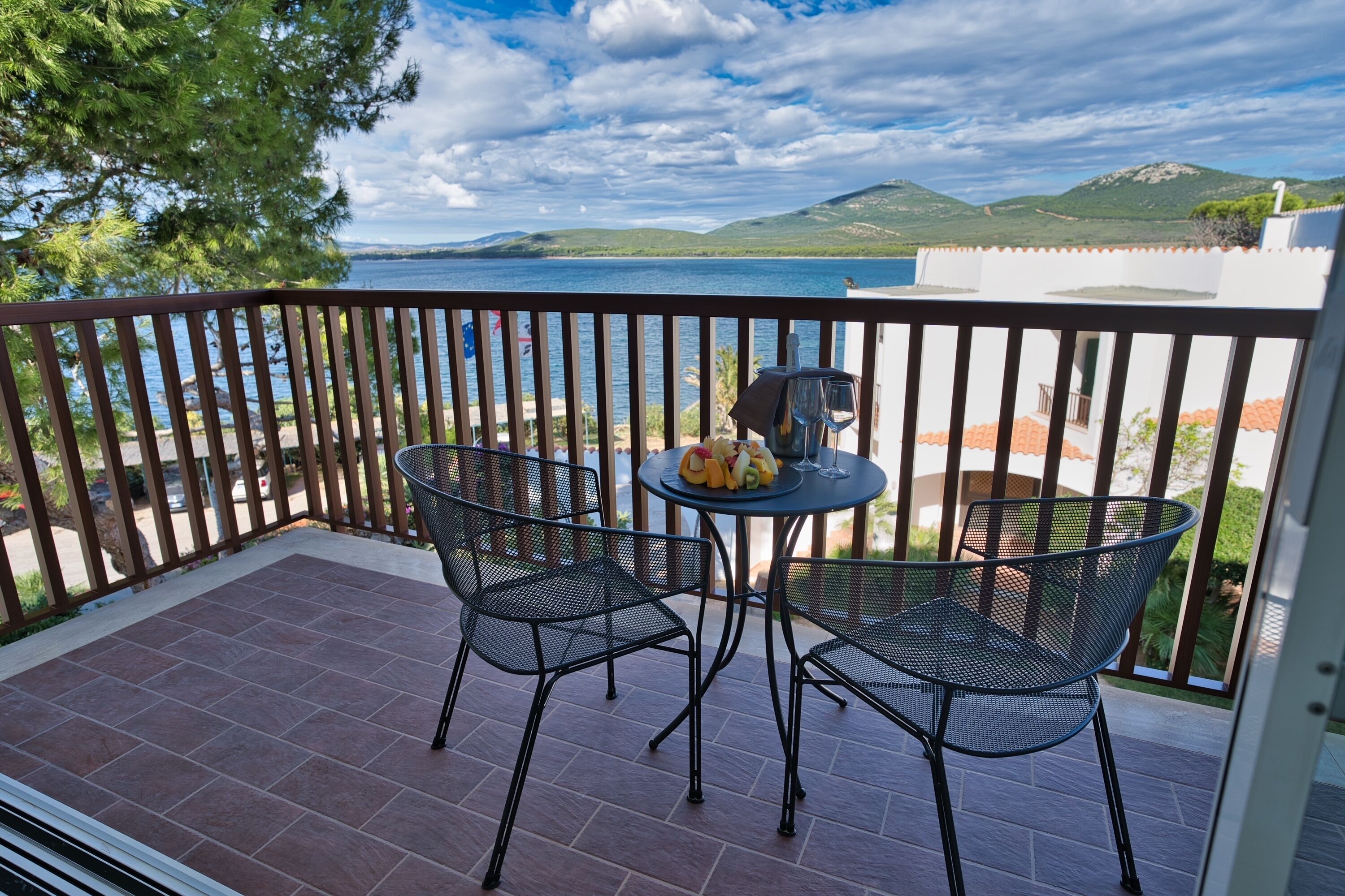 A balcony with two metal chairs and a small round table, overlooking a body of water and hills.