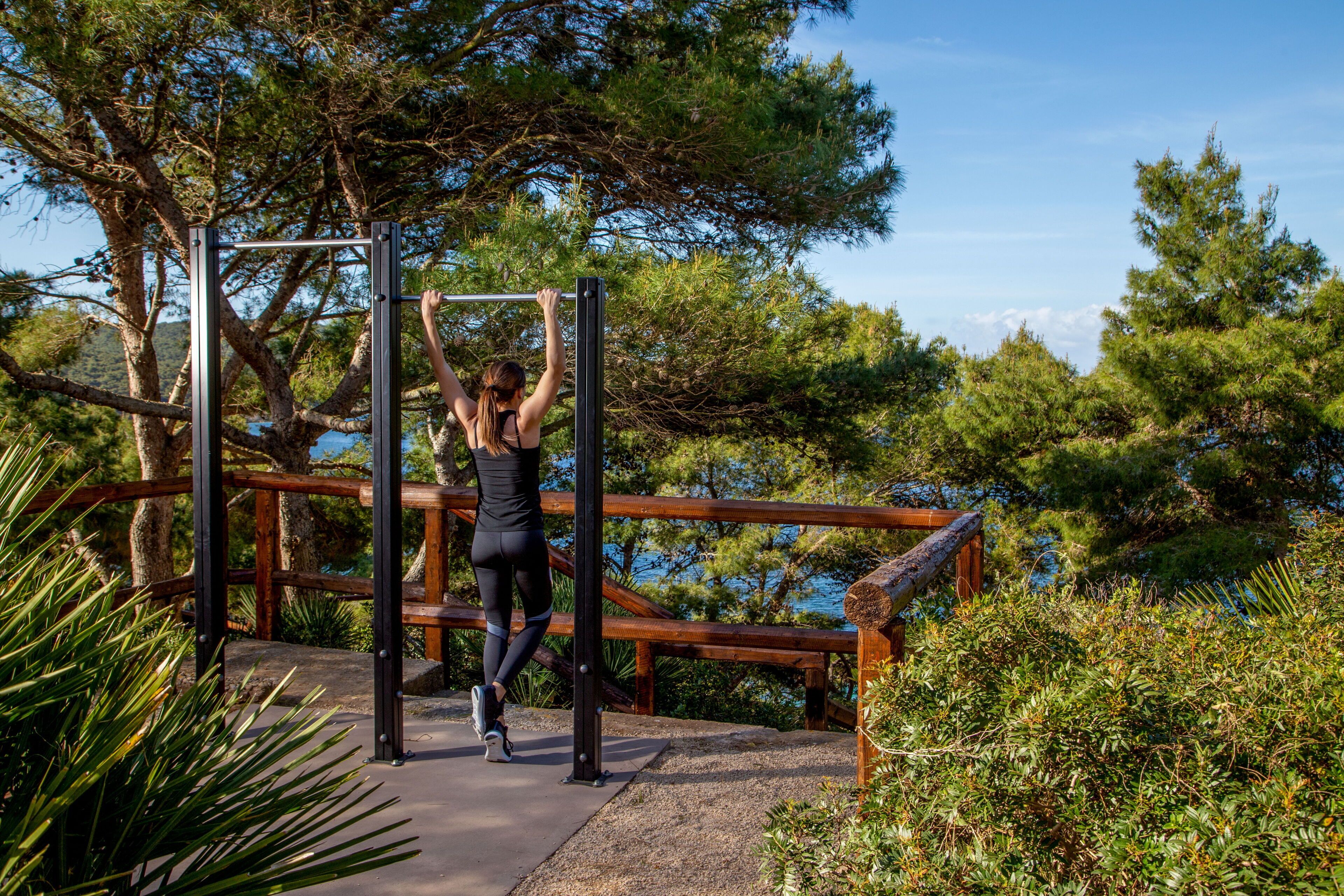A person performing pull-ups on a metal bar structure in a natural setting with trees and a wooden deck.