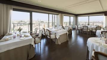 Un comedor con mesas dispuestas para el servicio, ventanas amplias que ofrecen vistas de la ciudad y un techo alto con iluminación empotrada.