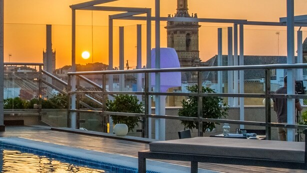 Une piscine sur le toit, entourée d’une balustrade en verre, offrant une vue sur le paysage urbain, notamment un beffroi, et un coucher de soleil en arrière-plan.