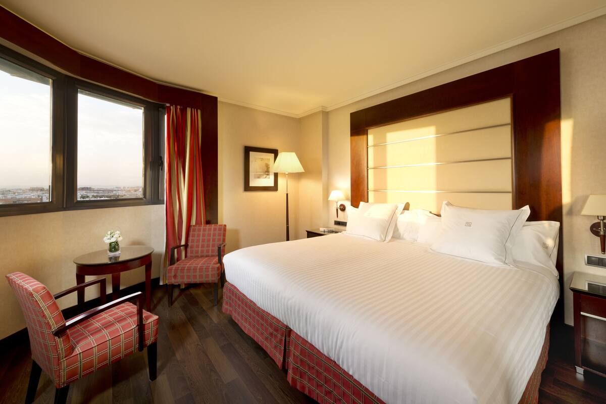 Habitación de hotel con una cama grande, un escritorio, dos sillas, una ventana con cortinas y vistas a la ciudad.