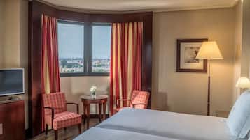 Una habitación de hotel con una cama grande, dos sillas, una mesita con un florero, un televisor y una lámpara.