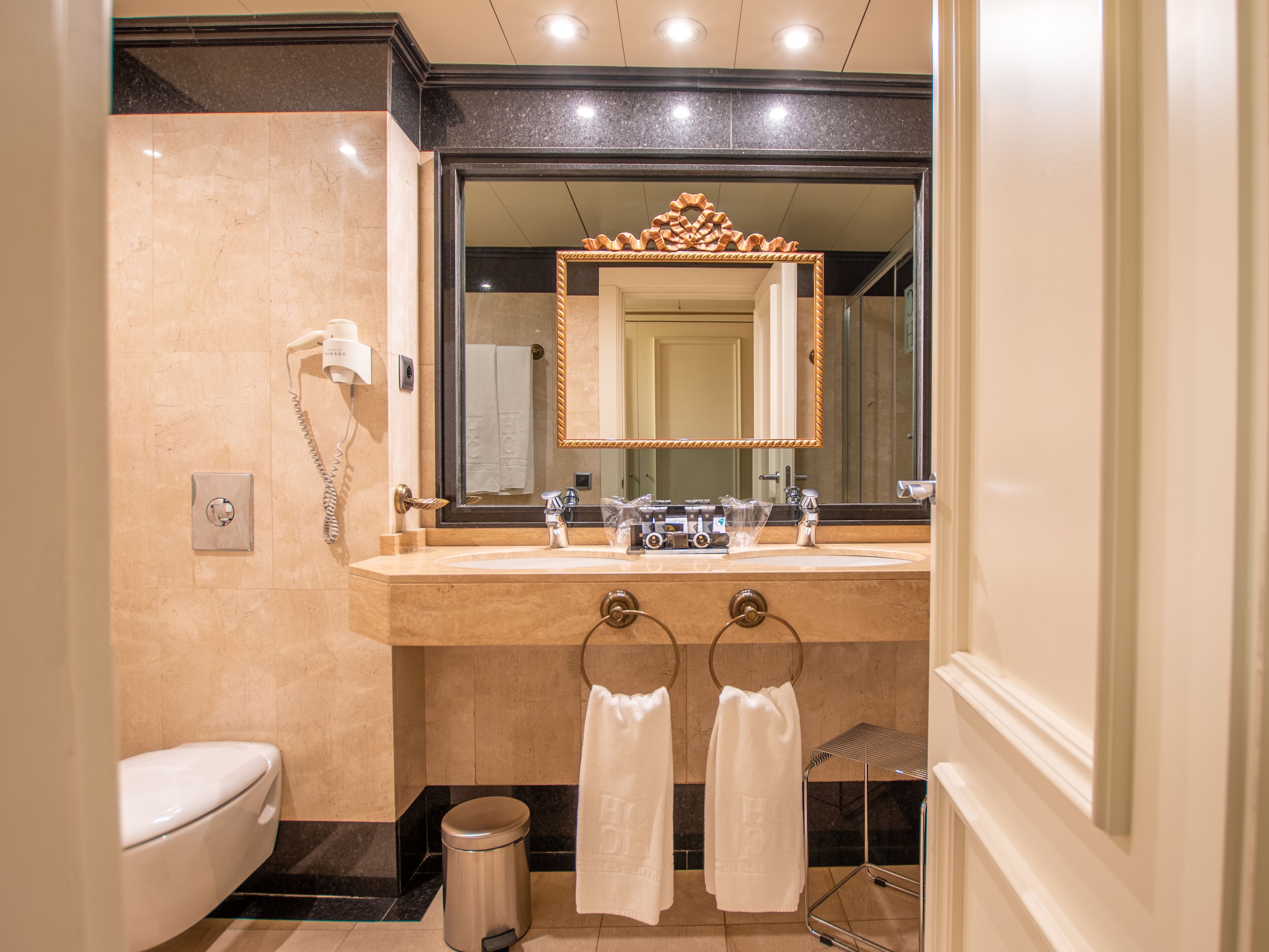 A modern bathroom with a marble sink, a large mirror, a hairdryer, and two towels hanging on hooks.