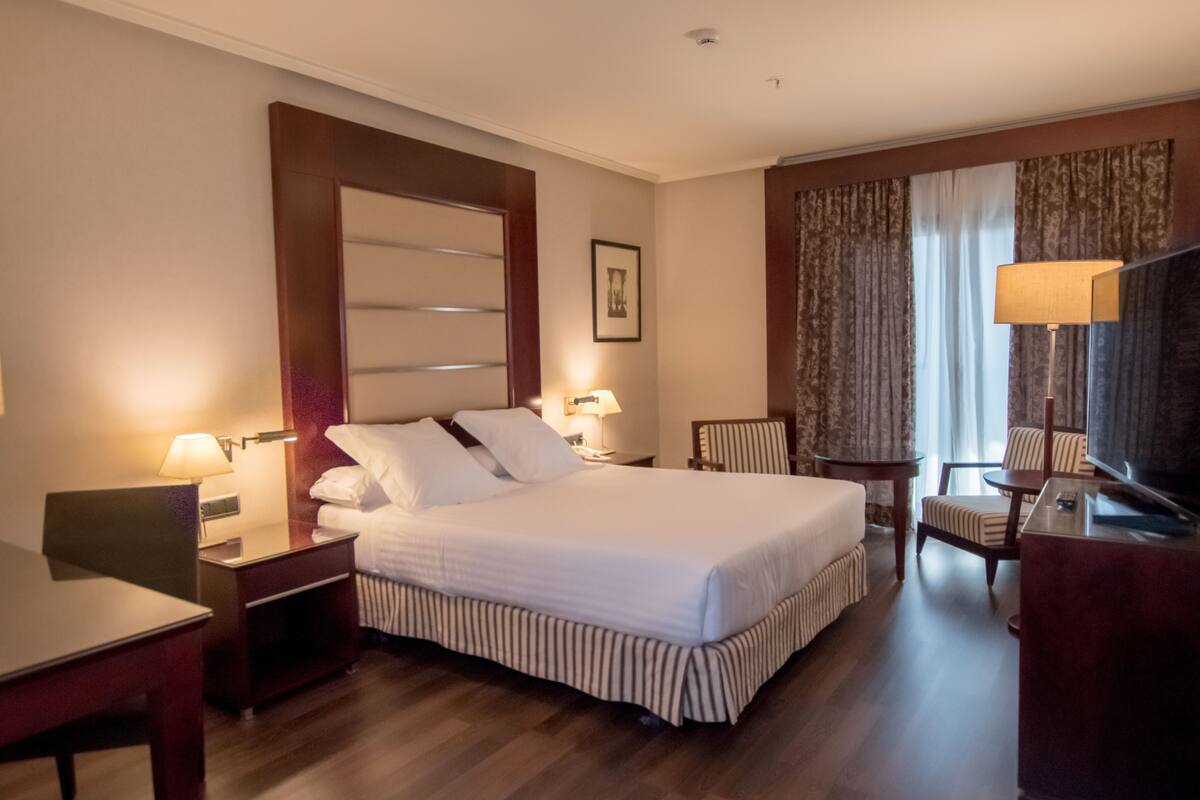 Habitación de hotel con una cama grande, mesitas de noche, un escritorio, una silla, un televisor y cortinas.