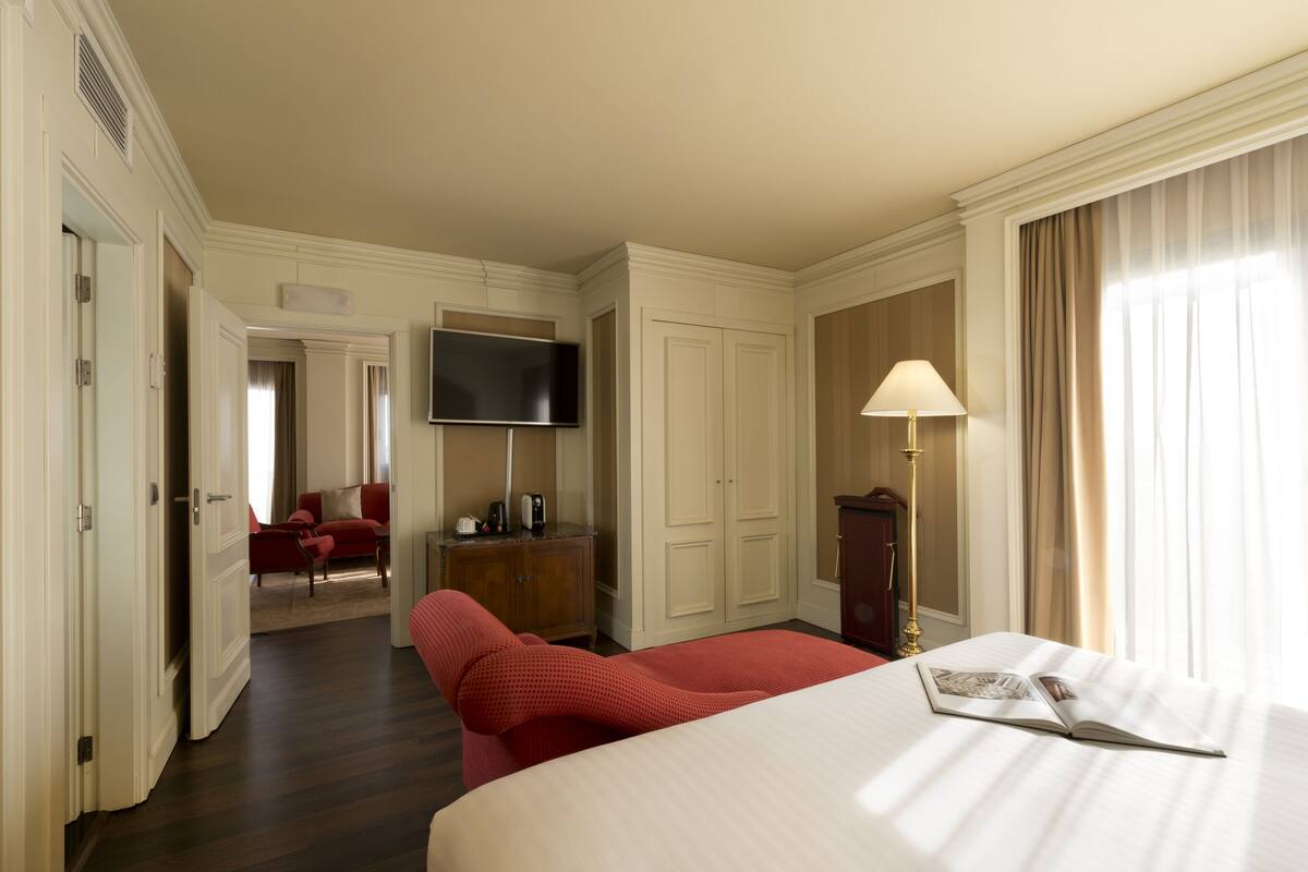 Una habitación de hotel con una cama grande, un televisor montado en la pared, un escritorio con lámpara y una silla roja.