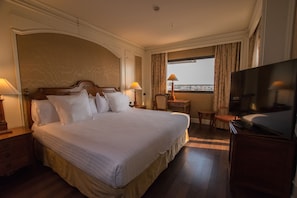A hotel room with a large bed, two bedside tables with lamps, a television, a small desk with a chair, and a window with curtains.