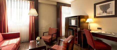 Habitación de hotel con un sofá rojo, dos sillones, una mesita con un jarrón, un televisor y un escritorio con una lámpara.