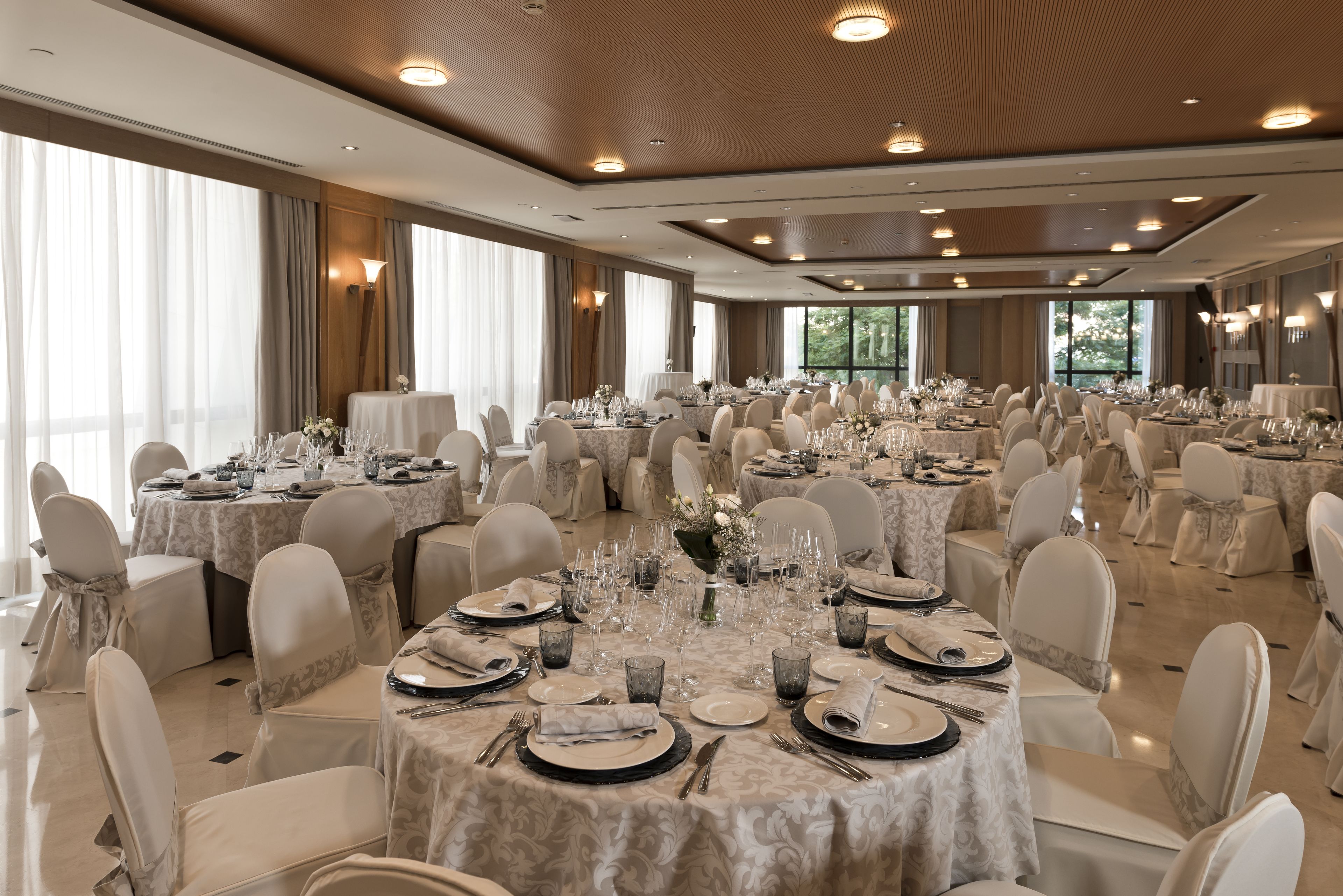 Un salón de banquetes con mesas redondas dispuestas para un evento formal, con sillas blancas y manteles.
