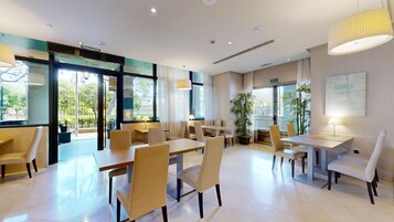 Un comedor moderno de hotel con mesas y sillas, ventanas amplias y plantas de interior.
