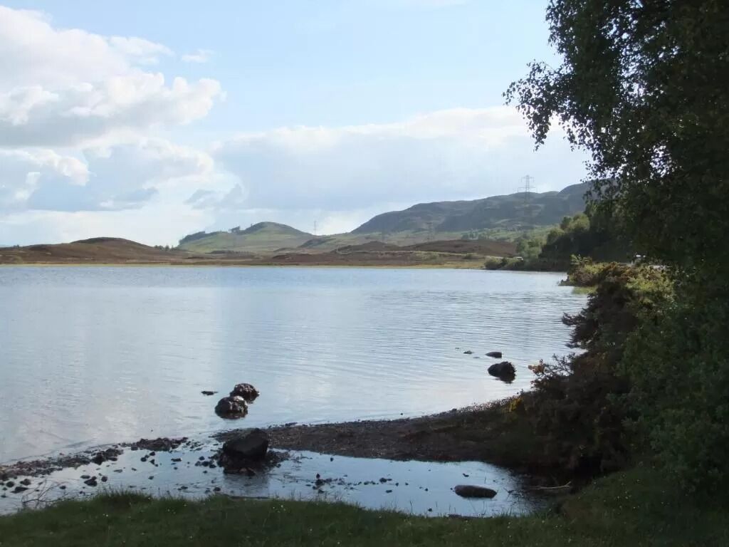 A calm lake with a grassy shore and hills in the background.