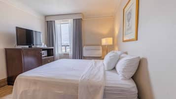 Ett hotellrum med en säng, en tv, ett skrivbord med en dator, en lampa och ett fönster med gardiner.