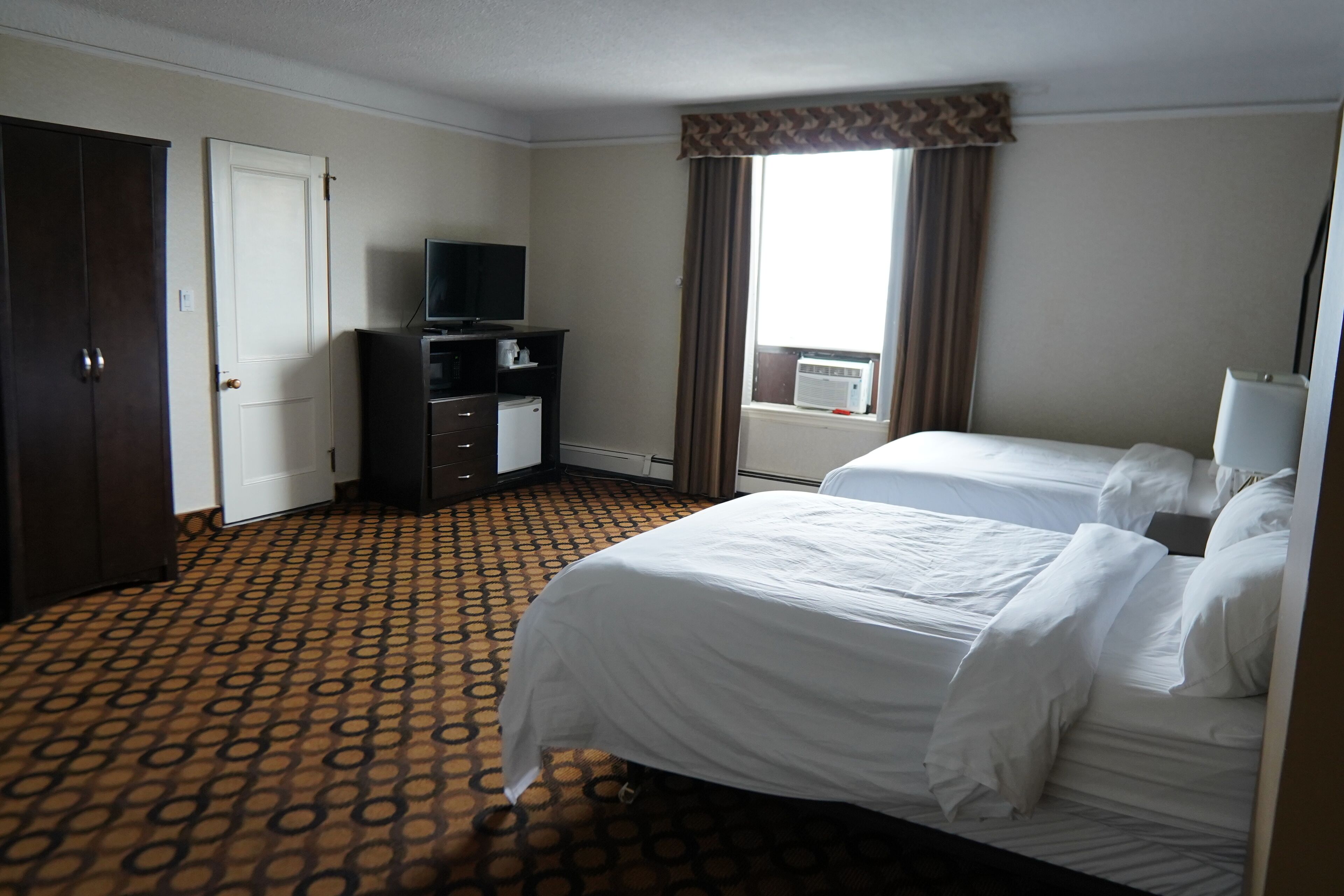 A hotel room with two beds, a TV, a desk, and a window with curtains.