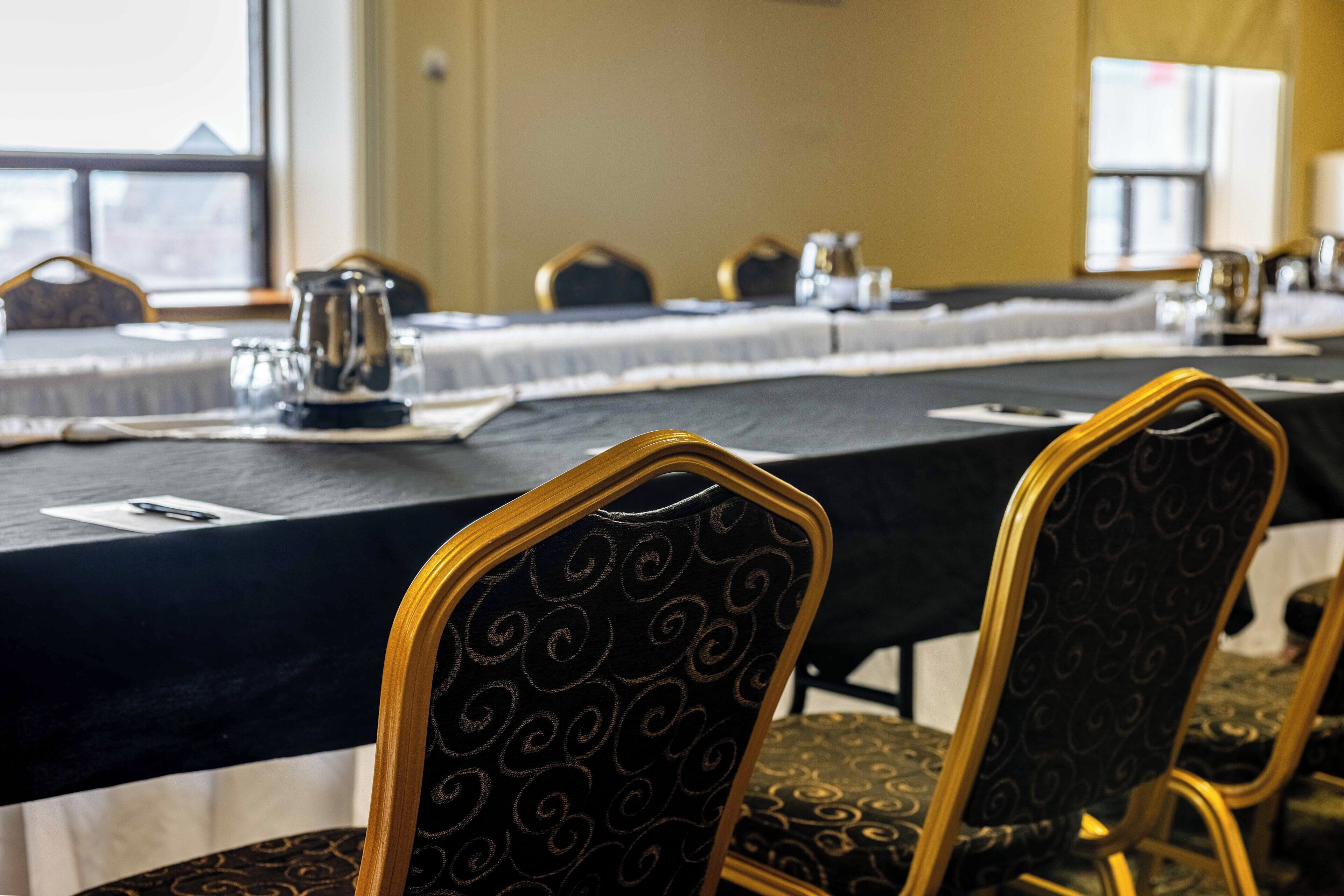 A conference room with a long table, chairs with patterned upholstery, and a window in the background.