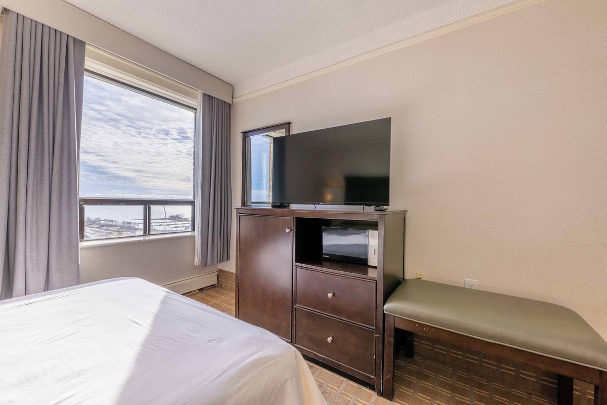 Una habitación de hotel con una cama, un televisor sobre un mueble de madera, una ventana con cortinas y un banco.