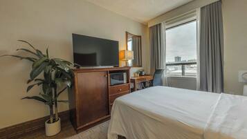 A hotel room with a bed, a TV, a desk, a chair, a potted plant, and a large window with curtains.