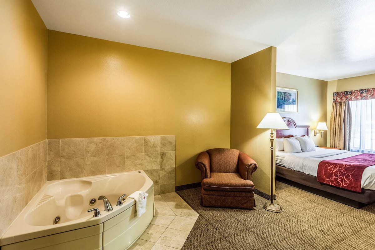 A hotel room with a bathtub, a chair, a bed, and a lamp.
