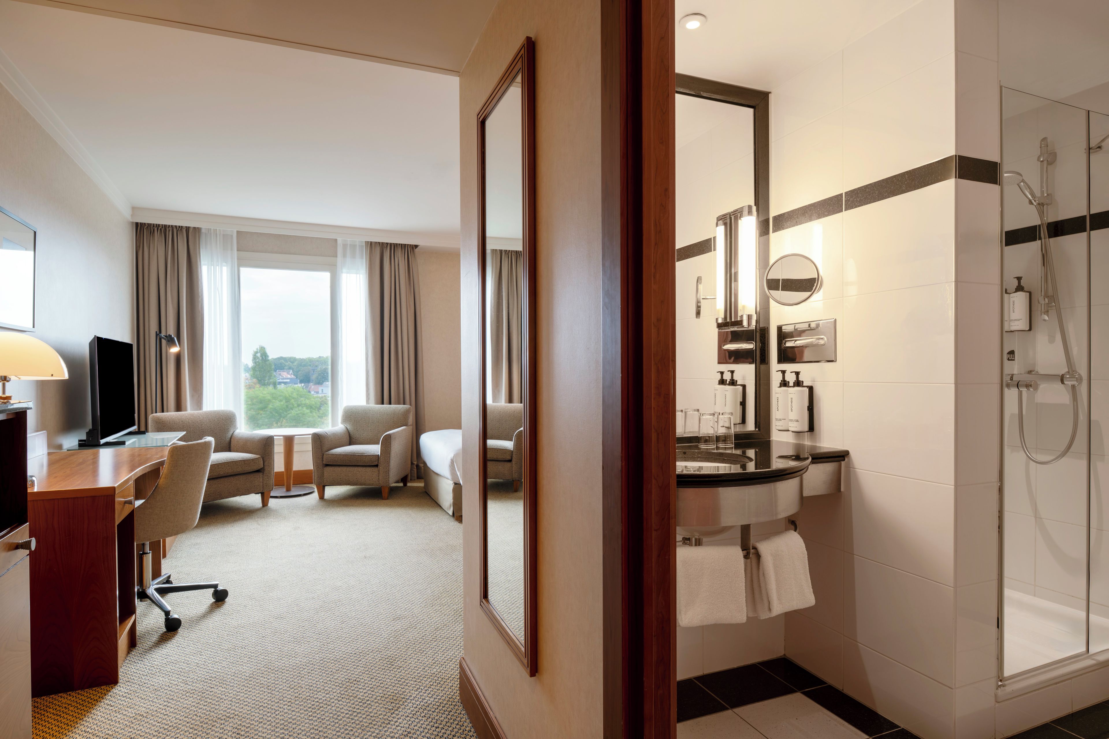 A hotel room with a desk, two armchairs, a bed, a large window with curtains, and a bathroom with a shower, sink, and mirror.
