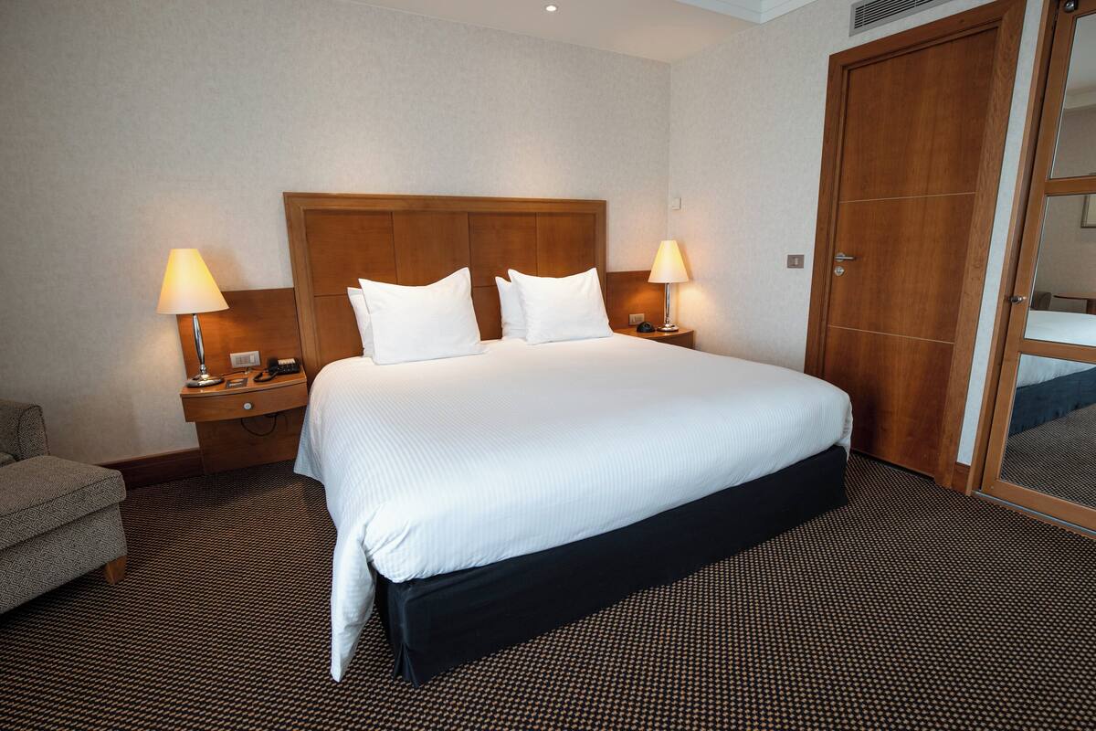 Une chambre d’hôtel avec un grand lit, deux tables de chevet avec des lampes, une tête de lit en bois, un lit recouvert d’une jupe noire et une moquette à motifs.