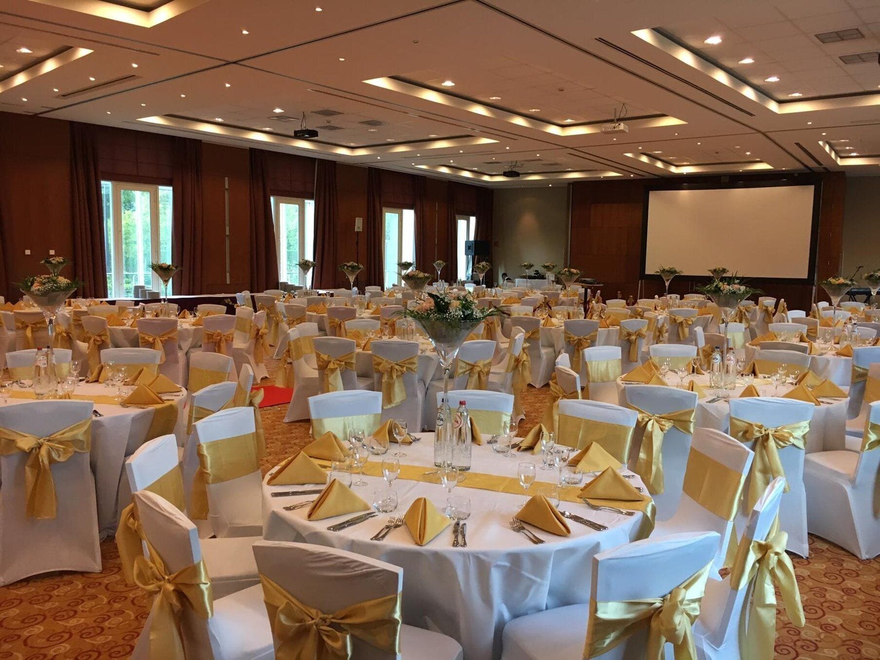 A banquet hall with round tables, white and gold chairs, and a projector screen.