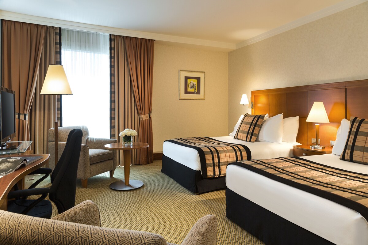 Ein Hotelzimmer mit zwei Betten, einem Schreibtisch, einem Stuhl und einem kleinen Tisch mit einer Lampe.