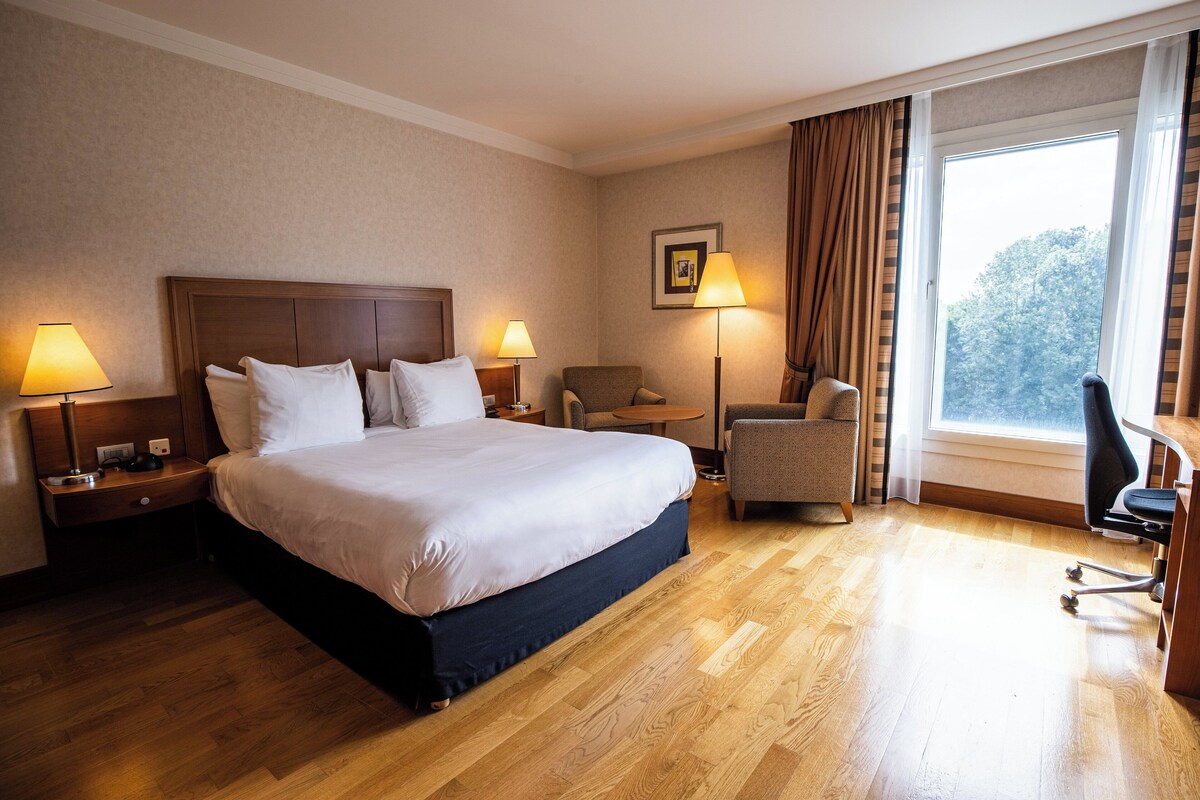 Ein Hotelzimmer mit einem großen Bett, zwei Nachttischen mit Lampen, einem Sessel, einem Schreibtisch und einem Fenster mit Vorhängen.