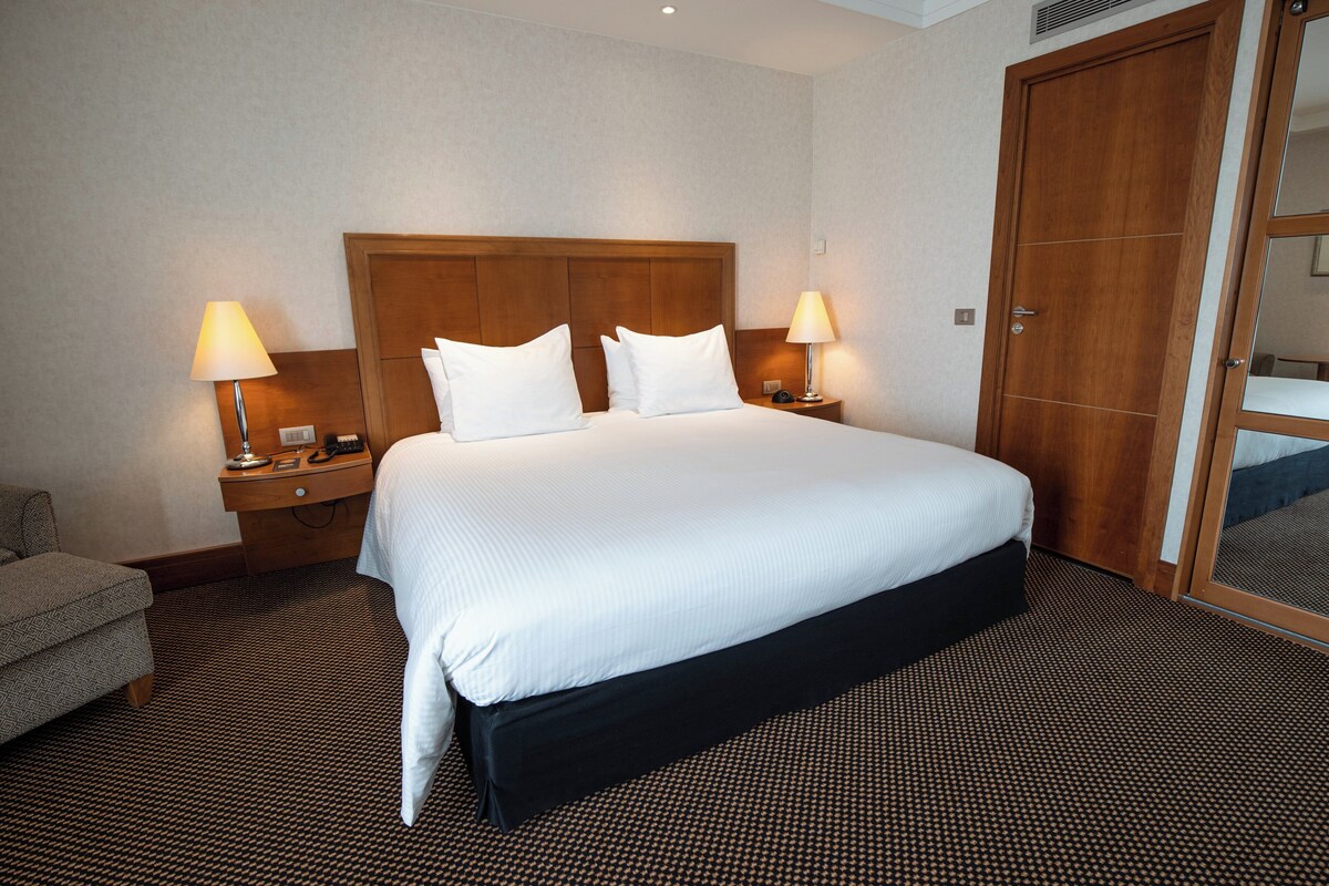 Ein Hotelzimmer mit einem großen Bett, zwei Nachttischen mit Lampen, einem Holz-Kopfteil und einem Bett mit schwarzem Betttuch sowie einem gemusterten Teppich.