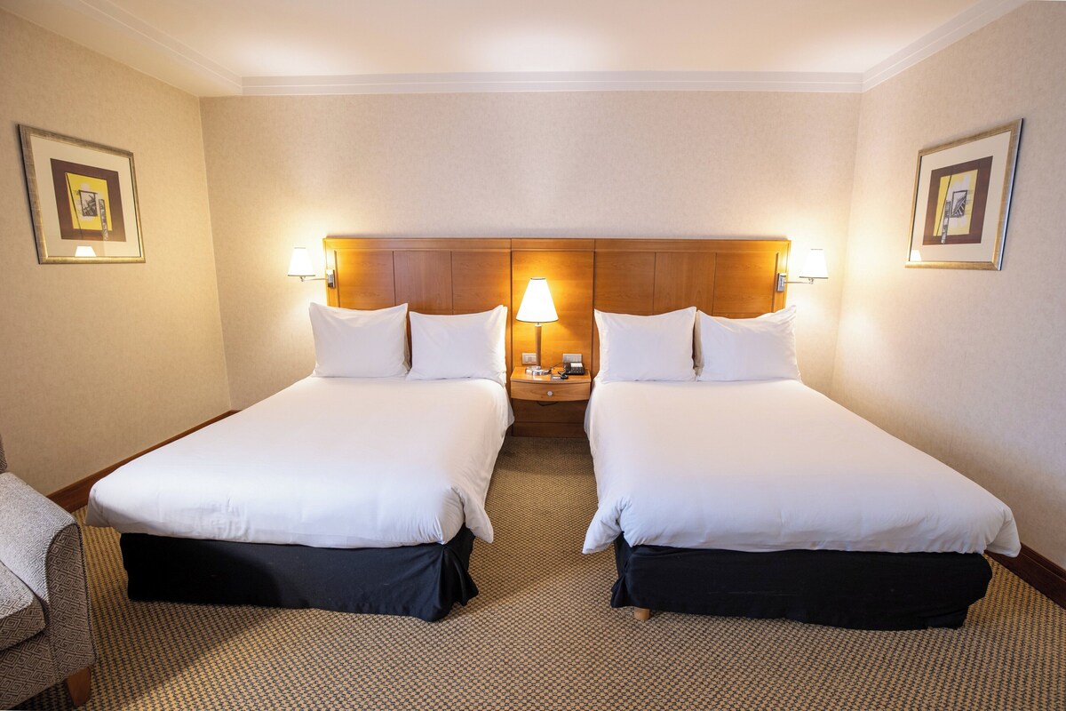 Ein Hotelzimmer mit zwei Betten, einem Nachttisch mit Lampe und gerahmten Bildern an der Wand.
