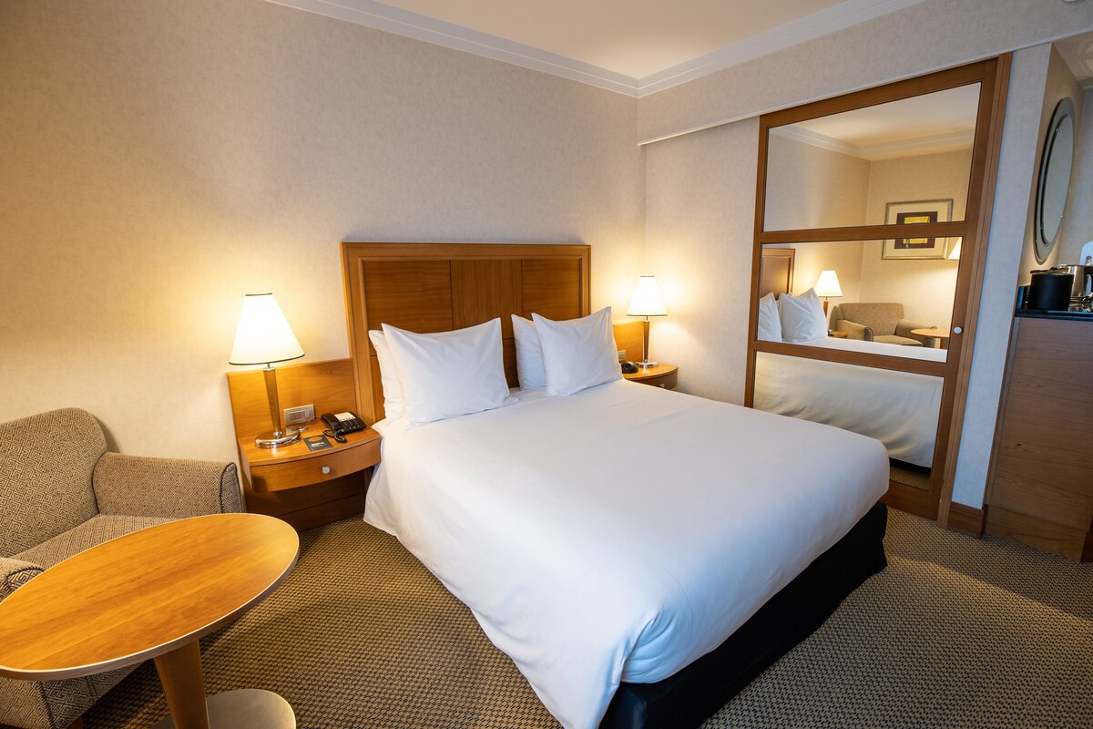 Ein Hotelzimmer mit einem großen Bett, zwei Nachttischen mit Lampen, einem kleinen runden Tisch, einer Couch und einem Spiegel.