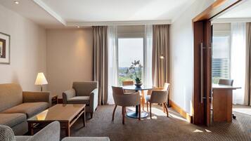 Een hotelkamer met een bank, fauteuil, eettafel en stoelen. Er is een raam met uitzicht op gebouwen en een deur aan de rechterkant.