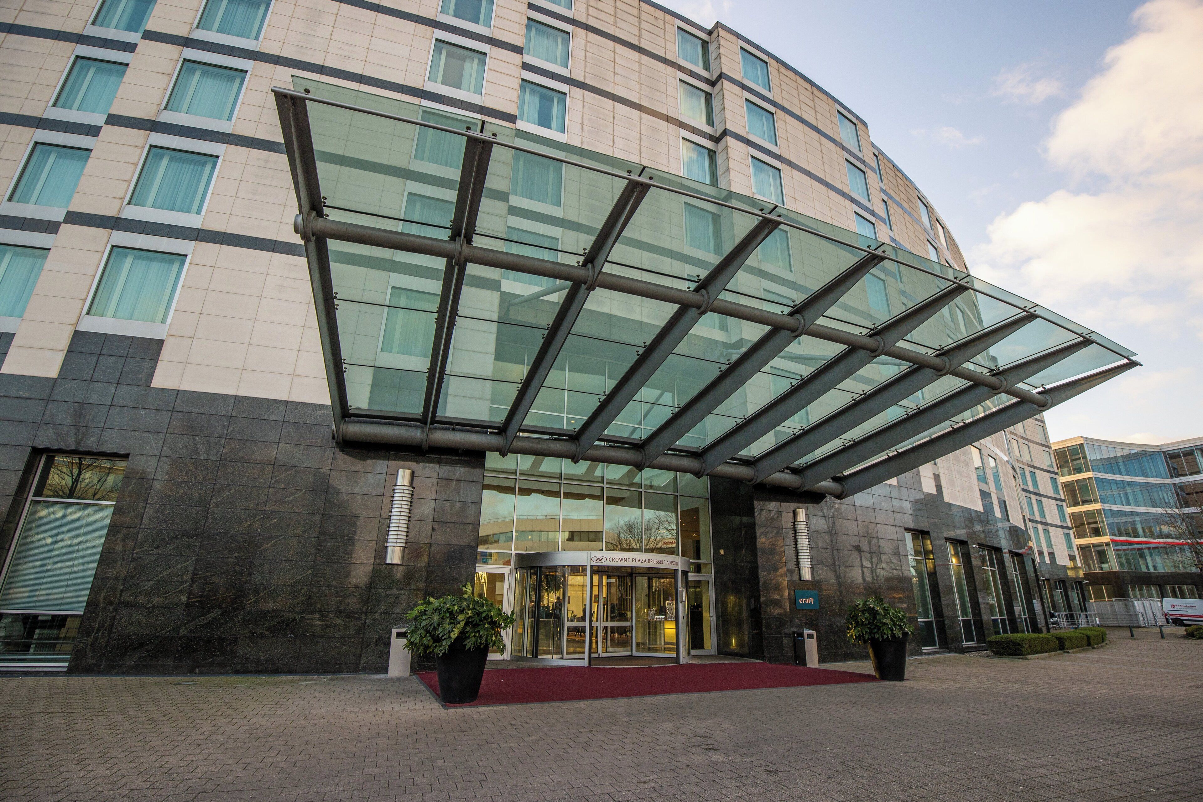 A modern hotel entrance with a glass canopy, a red carpet, and potted plants.