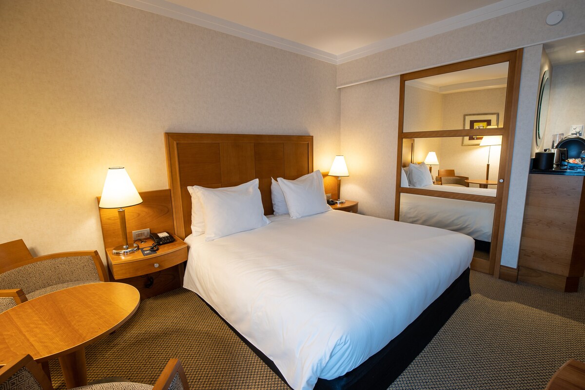 Ein Hotelzimmer mit einem großen Bett, zwei Nachttischen mit Lampen, einem Holz-Kopfteil, einem runden Tisch und einem Spiegel.