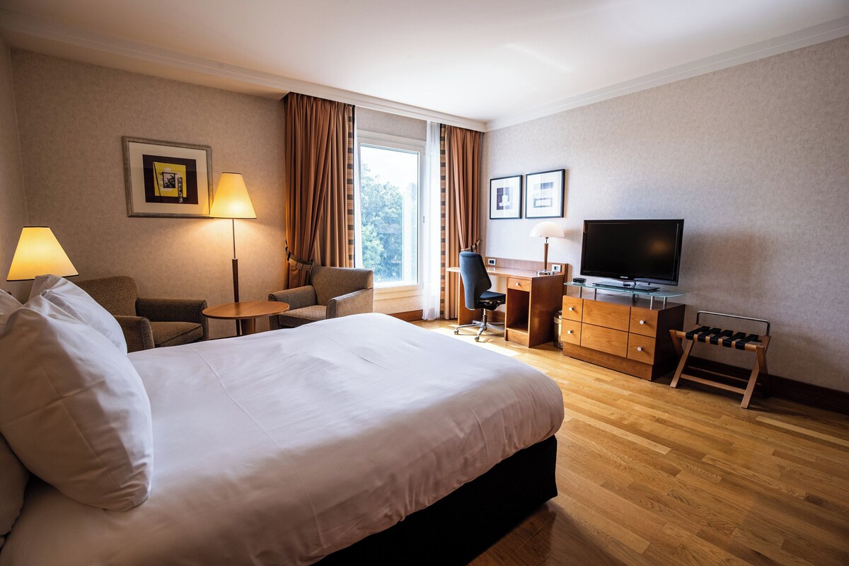 Ein Hotelzimmer mit einem großen Bett, einem Schreibtisch mit Computer, einem Fernseher und einer Sitzecke mit Stühlen und einem Tisch.