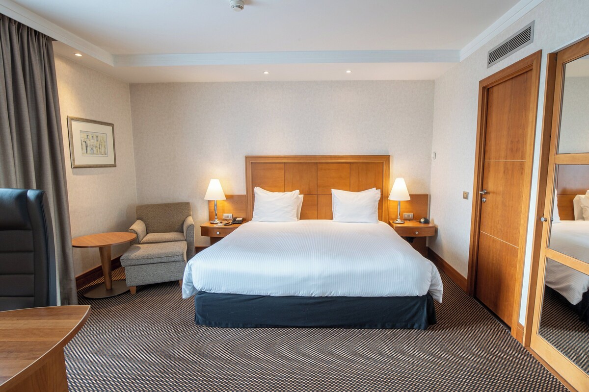 Ein Hotelzimmer mit einem großen Bett, zwei Nachttischen mit Lampen, einem Sessel, einem Schreibtisch und einem Kleiderschrank.