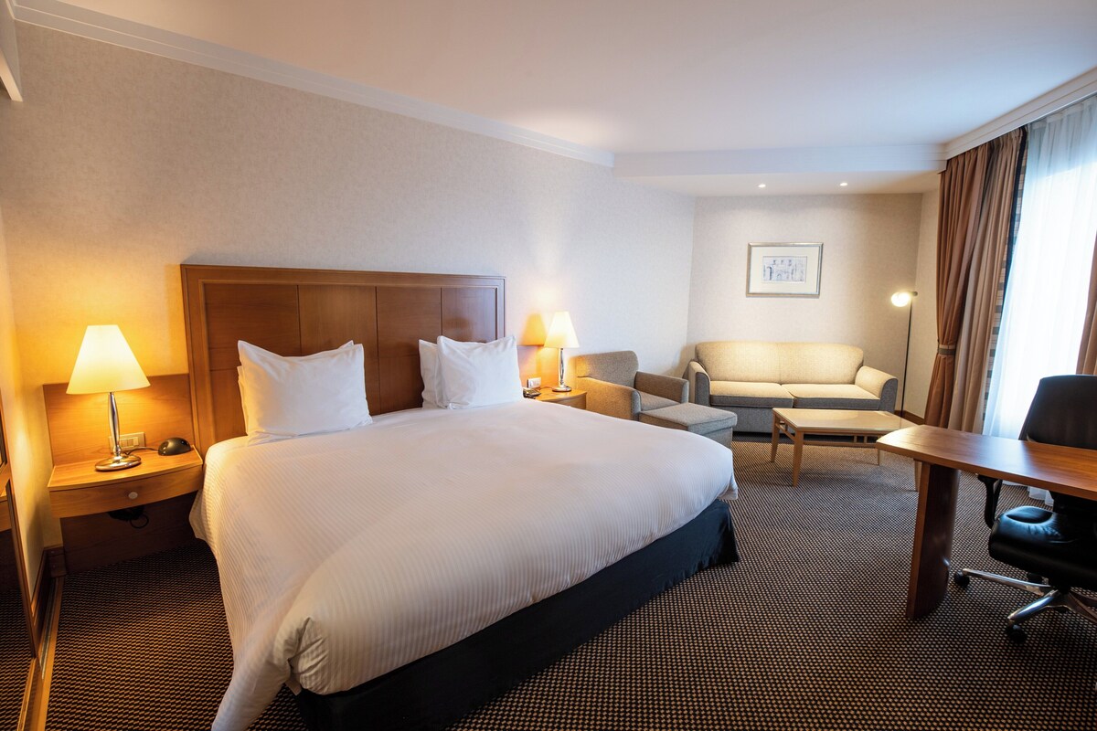 Ein Hotelzimmer mit einem großen Bett, zwei Nachttischen inklusive Lampen, einem Sofa, einem kleinen Tisch und einem Schreibtisch mit Stuhl.