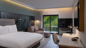 Ein modernes Hotelzimmer mit einem großen Bett, einem Schreibtisch, einem Sessel und einem Balkon mit Blick auf Grünflächen.